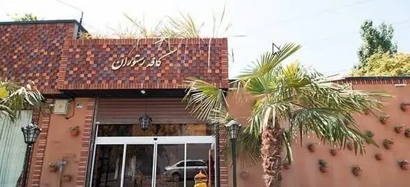 تصویری از نمای خارجی شیک کافی شاپ شاندیز اعیانی، شامل تابلوی کافه رستوران، درب ورودی شیشه‌ای، نخل‌های زیبا و دیوار تزئین شده با گلدان.