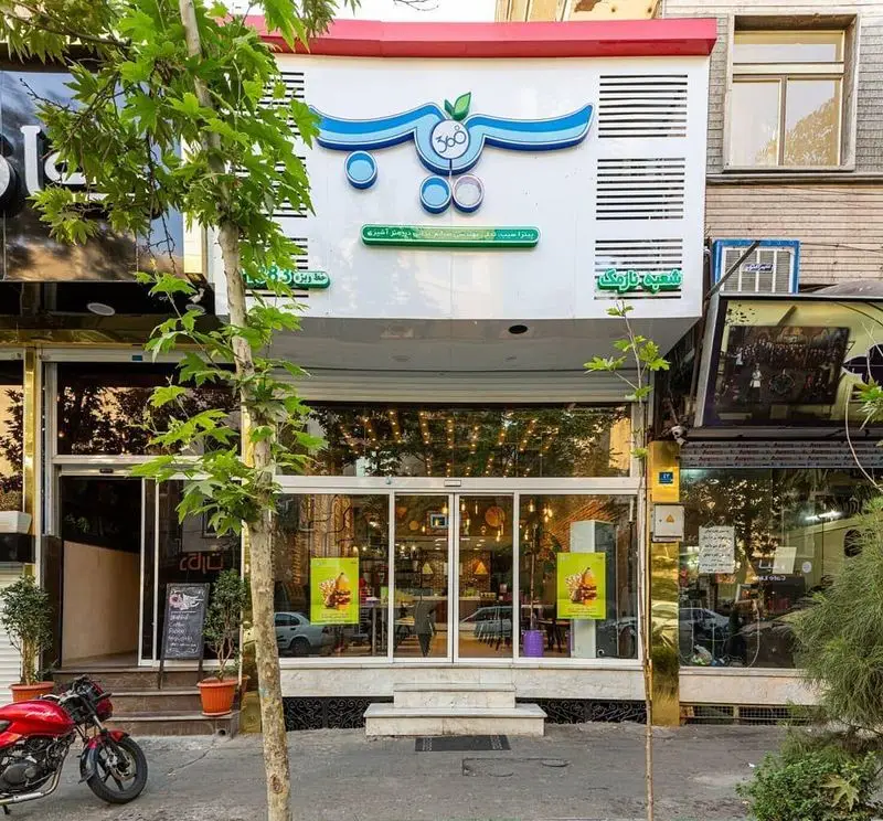 نمای بیرونی جذاب فست فود سیب ۳۶۰ با تابلوی اصلی برند، ویترین‌های شیشه‌ای بزرگ، درخت کنار خیابان و محیط شهری اطراف.