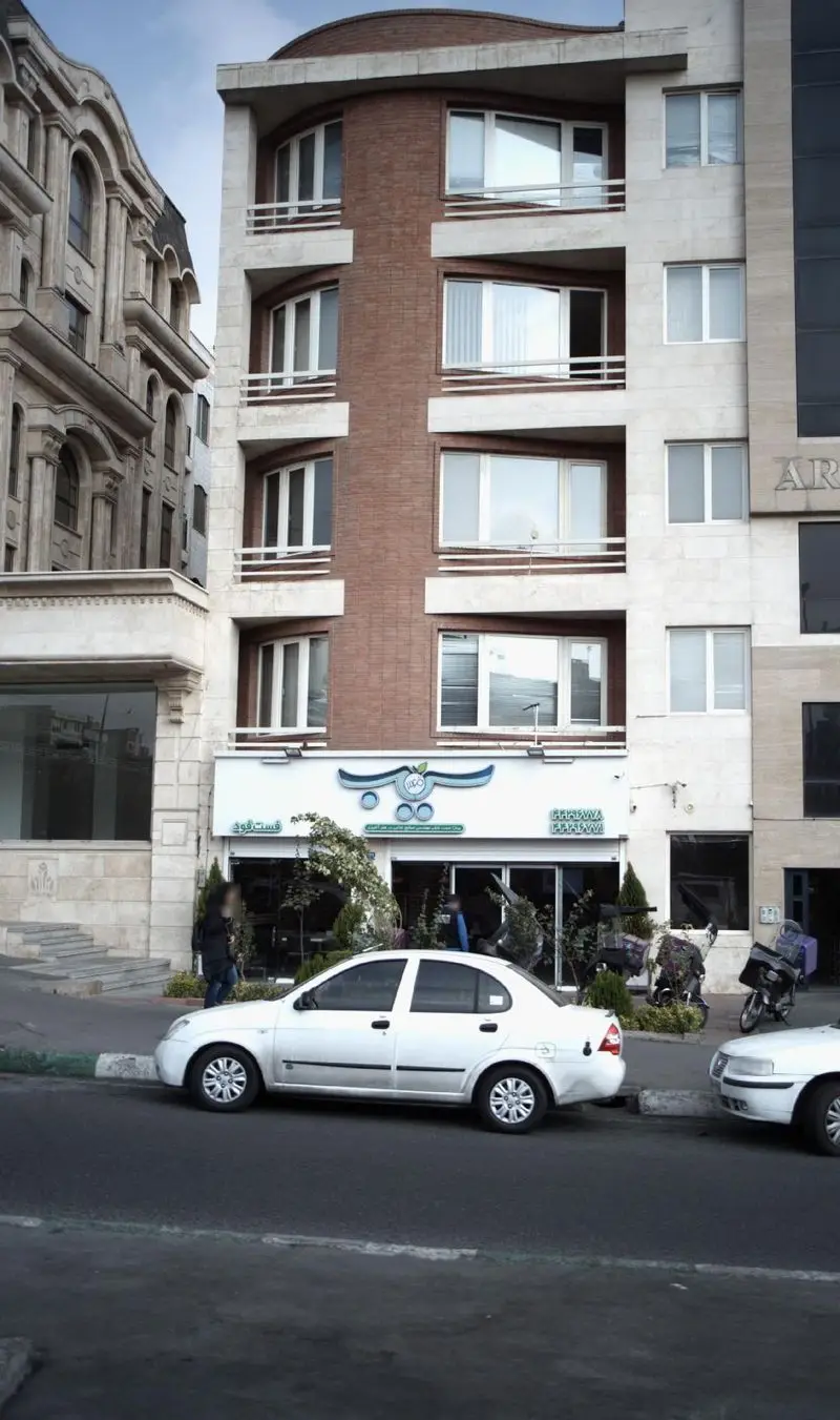 تصویری از نمای بیرونی فست فود سیب ۳۶۰ با تابلو آبی رنگ و لوگوی سیب، همراه با ورودی شیشه‌ای، گل و گیاه و ماشین پارک شده در خیابان