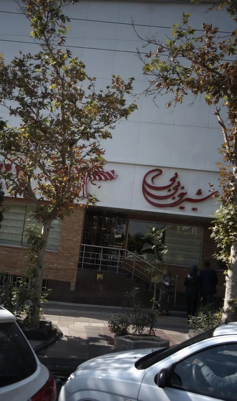 تصویری زیبا از ورودی شیرینی بی بی، یک قنادی معروف با تابلوی قرمز رنگ، همراه با درختان سبز و نمای خیابان در روز روشن.