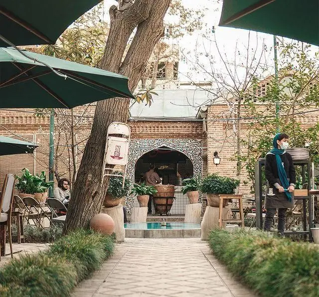 کافه روحی (باغ نگارستان): حیاطی دلنشین با معماری سنتی، حوضچه آبی، درخت کهنسال، میز و صندلی فضای باز و پرسنل.