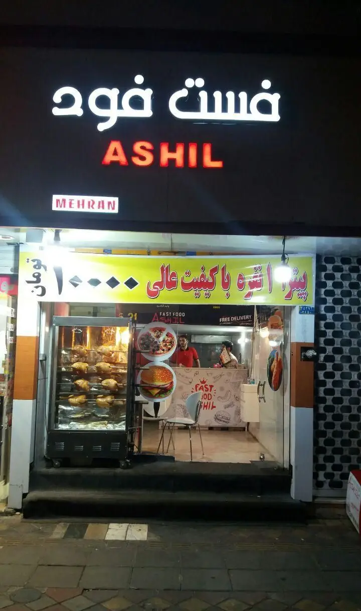 نمای بیرونی فست فود پیتزا آشیل (Ashil) با تابلو روشن در شب، ارائه دهنده پیتزا با کیفیت عالی، مرغ بریان و برگر.