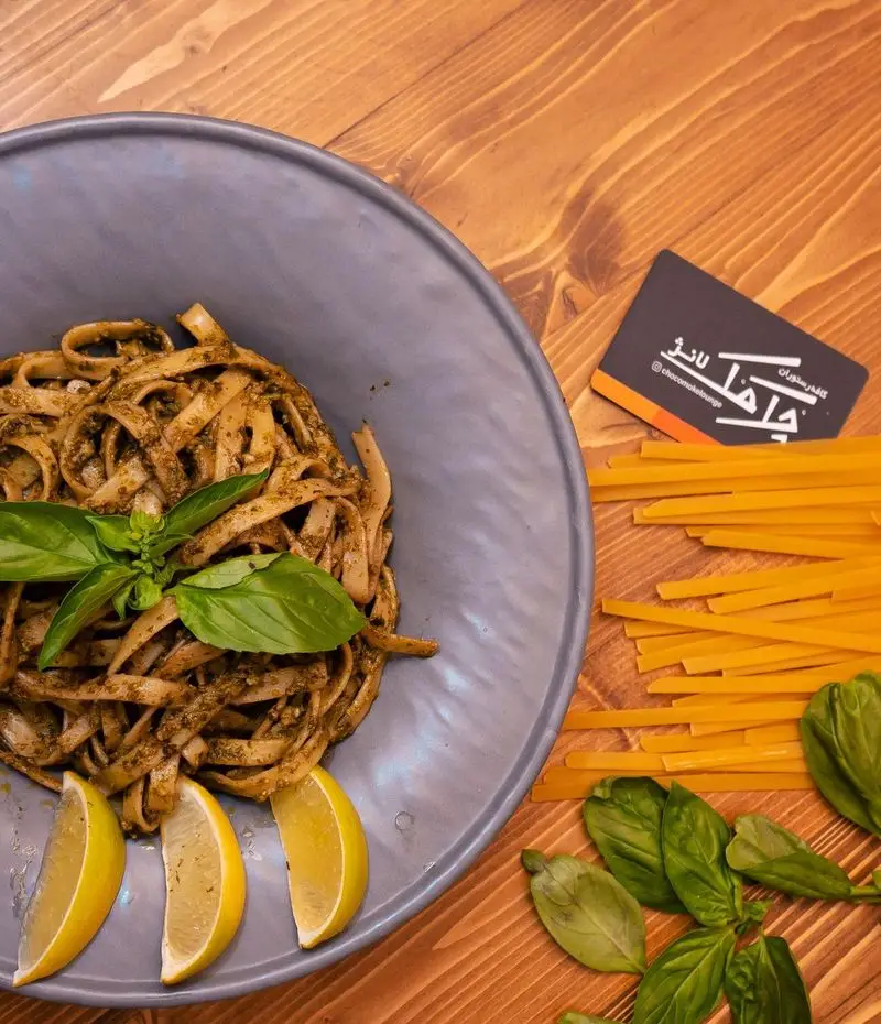 پاستا پستو فتوچینی خوشمزه کافه رستوران چک مک لانژ با تزئین ریحان تازه و برش‌های لیمو، همراه با پاستای خام روی میز چوبی.