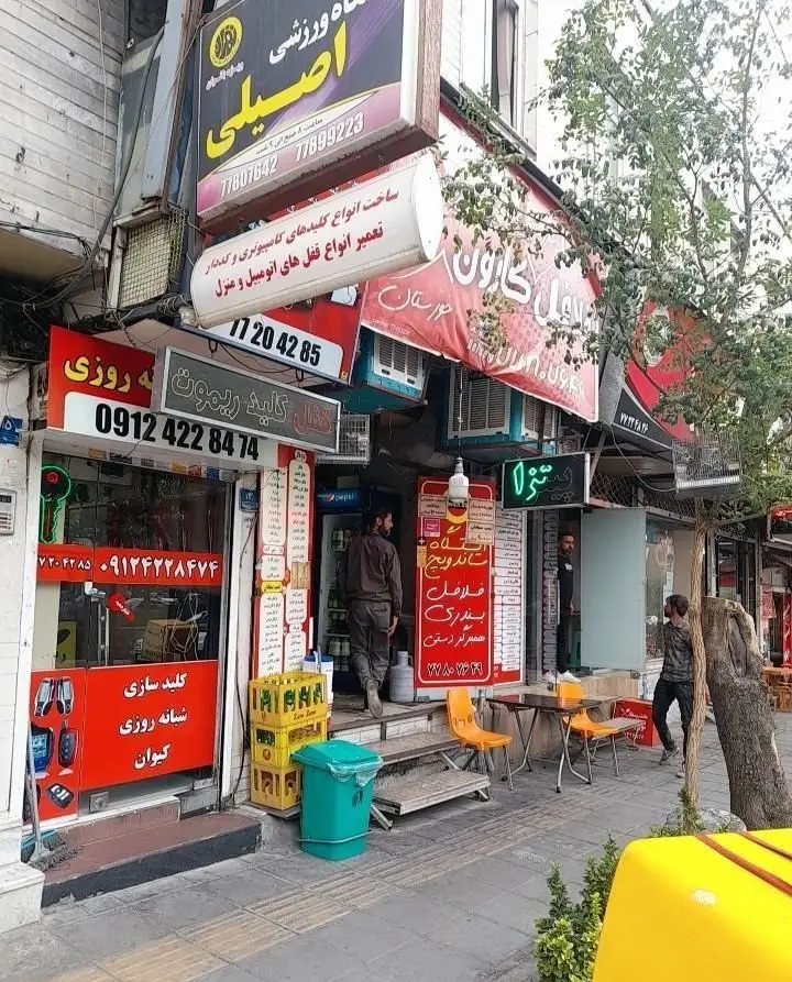 فست فود کارون خوزستان با تابلوی قرمز و میز و صندلی‌های بیرون، در کنار مغازه کلیدسازی و سایر کسب و کارها در یک خیابان پر جنب و جوش.