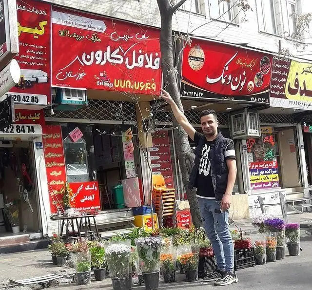 نمای بیرونی فست فود و فلافل کارون خوزستان با تابلوی روشن. مردی به نام مغازه اشاره می‌کند. خدمات تحویل و سفارش غذا.