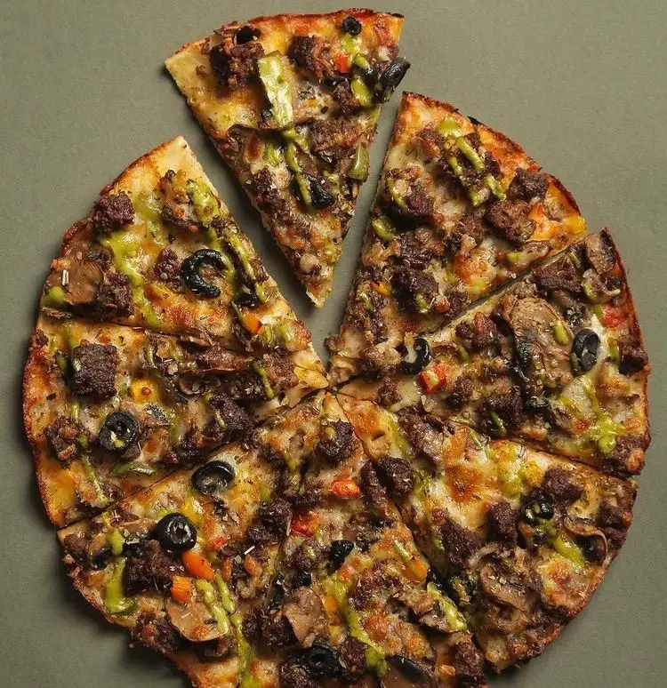 پیتزای خوشمزه فست فود بولکی با تکه‌های گوشت، قارچ، زیتون سیاه و سس سبز، یک غذای لذیذ برای تجربه طعمی بی‌نظیر.
