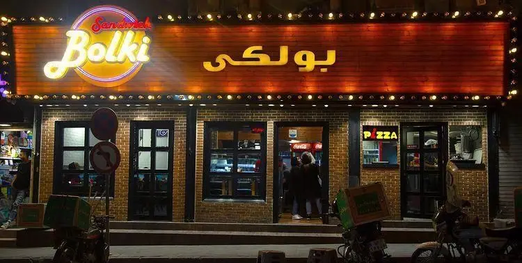 نمای شبانه فست فود بولکی، رستورانی محبوب برای انواع ساندویچ و پیتزا، با تابلوی نئون درخشان و موتورهای دلیوری آماده سرویس دهی.