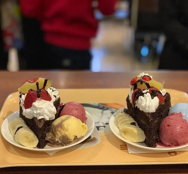 دو برش کیک شکلاتی با خامه، توت فرنگی، آناناس و موز، همراه با اسکوپ های بستنی لذیذ. دسرهای ویژه و خوشمزه کافی شاپ گل یخ.
