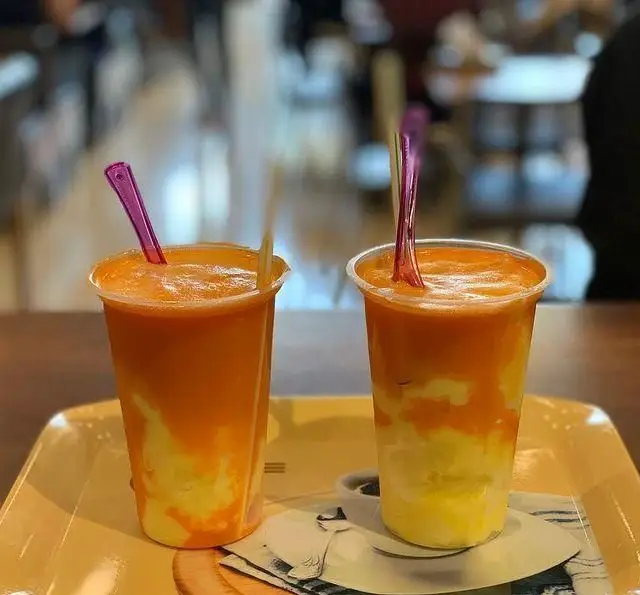 لذت نوشیدنی‌های تابستانی در کافی شاپ گل یخ: دو لیوان اسموتی دولایه میوه‌ای خنک با رنگ‌های شاد زرد و نارنجی، تجربه‌ای بی‌نظیر.