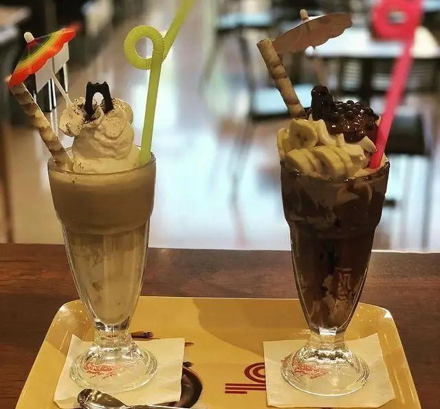 تصویری دلنشین از دو میلک شیک رنگارنگ و خوش‌طعم در کافی شاپ گل یخ، شامل میلک شیک قهوه و شکلات موز، با تزیینات جذاب.