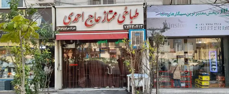 مغازه طباخی ممتاز حاج احمدی با تابلوی برجسته قرمز، ارائه دهنده کله پاچه و سیراب شیردان، در یک نمای خیابانی با درختان.