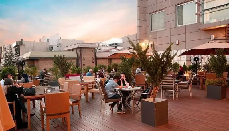 فضای باز و دلنشین کافی شاپ دلچی، با مشتریانی که در حال لذت بردن از نوشیدنی‌ها و دورهمی دوستانه در یک عصر زیبا هستند.