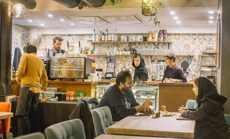 فضای داخلی دنج و دلنشین کافی شاپ دلچی با مشتریانی که در حال نوشیدن قهوه و چای هستند و کارکنان آماده خدمت‌رسانی.