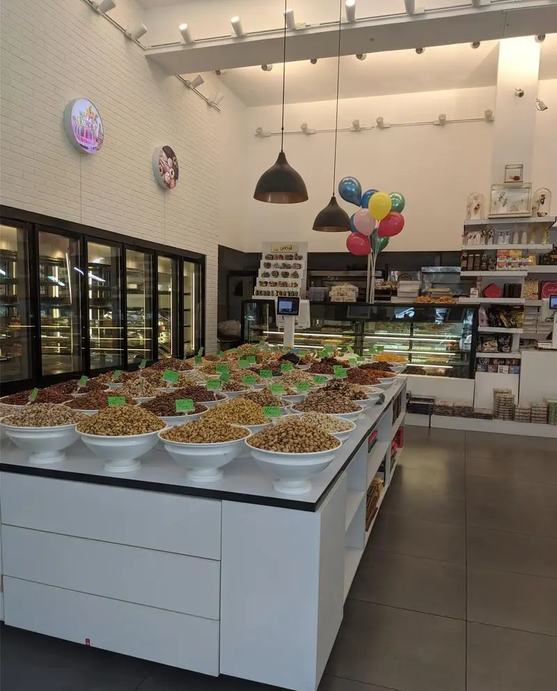 فروشگاه بزرگ نان و شیرینی لادن غرب، نمایش آجیل و خشکبار متنوع در ظروف سفید، قنادی شیک با ویترین شیرینی‌ها و دکور مدرن.