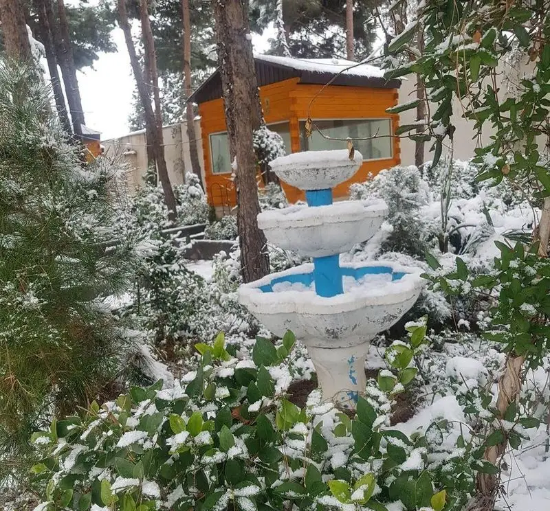 تصویری زیبا از باغ رستوران بهشت پنهان در فصل زمستان، با فواره سه طبقه برفی و پوشش گیاهی سبز. منظره‌ای آرامش‌بخش.