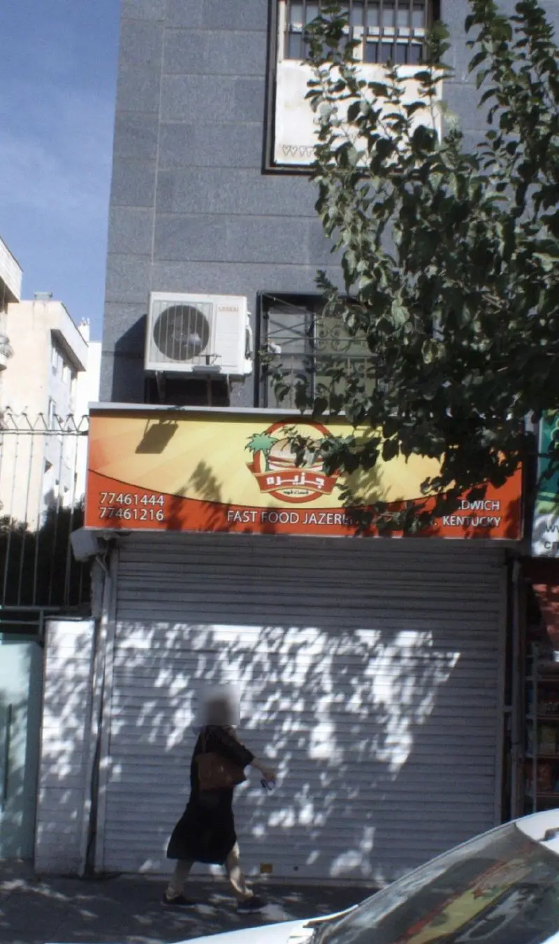 نمای بیرونی فست فود جزیره با تابلوی نارنجی-قرمز، شامل شماره های تماس 77461444 و 77461216. ارائه دهنده ساندویچ و کنتاکی. فروشگاه بسته.