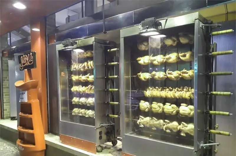 مرغ‌های بریان آبدار و طلایی در حال پخت داخل دستگاه‌های روتاری فست فود فدک، آماده سرو برای مشتریان. تابلوی فلافل در کنار.