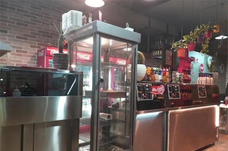 تصویری از داخل فست فود فدک: پیشخوان‌های استیل، یخچال نوشابه کوکاکولا، منوهای متنوع و فضای تمیز و دکوراسیون مدرن.