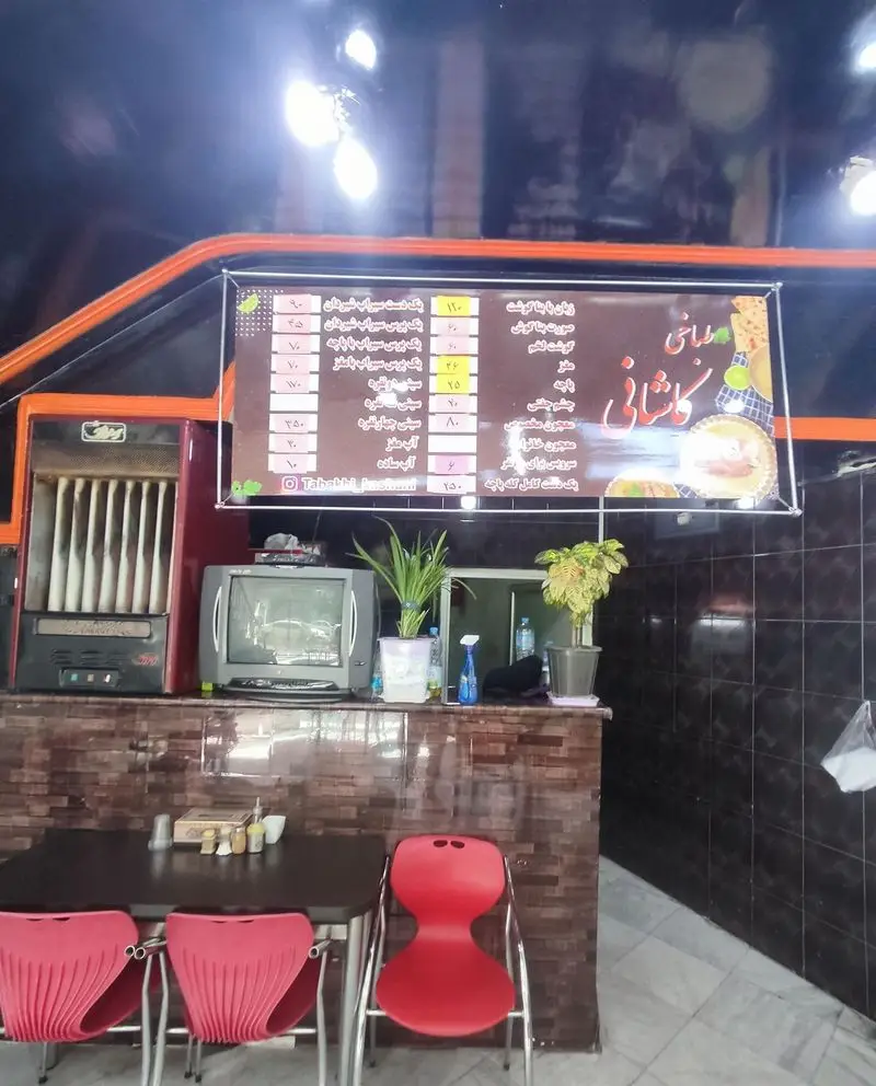 فضای داخلی طباخی کاشانی با منوی کله پاچه، میز و صندلی‌های قرمز و کانتر پذیرایی. محیطی سنتی و گرم برای مشتریان.