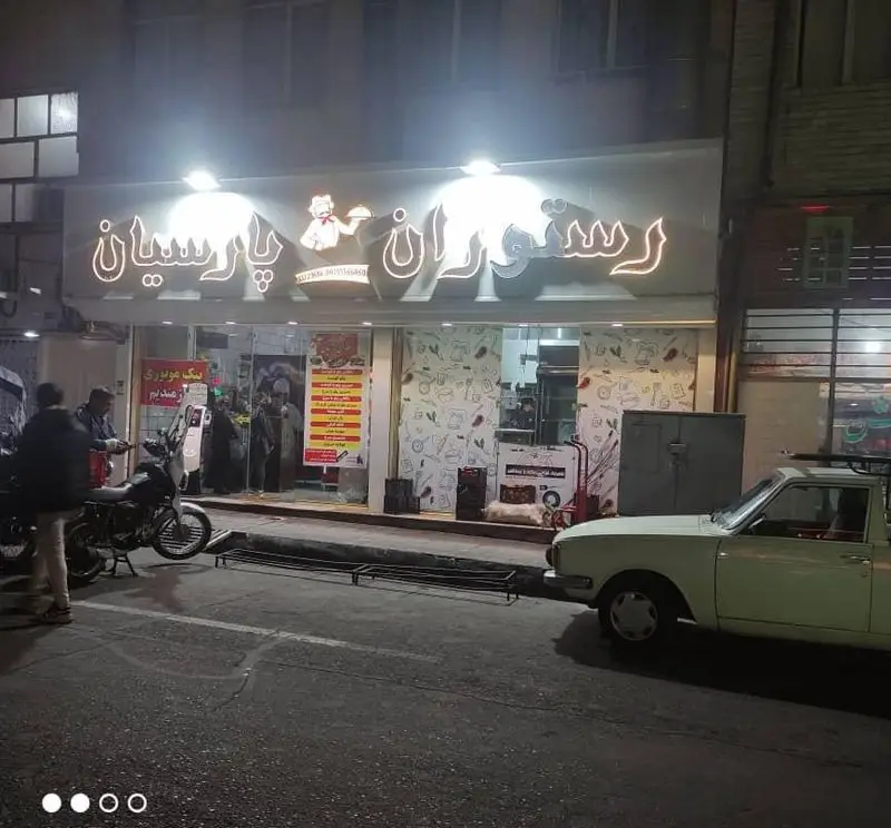 نمای شبانه رستوران پارسیان رضایی با تابلوی نورانی، لوگوی آشپز، ورودی شیشه‌ای، مشتریان، موتورسیکلت و خودرو پارک شده.