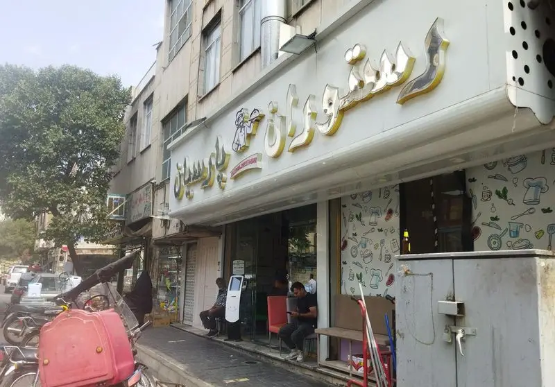 عکس خیابانی از رستوران پارسیان رضایی، با تابلوی روشن، طرح‌های آشپزی روی پنجره و مشتریان نشسته جلوی ورودی در روز آفتابی