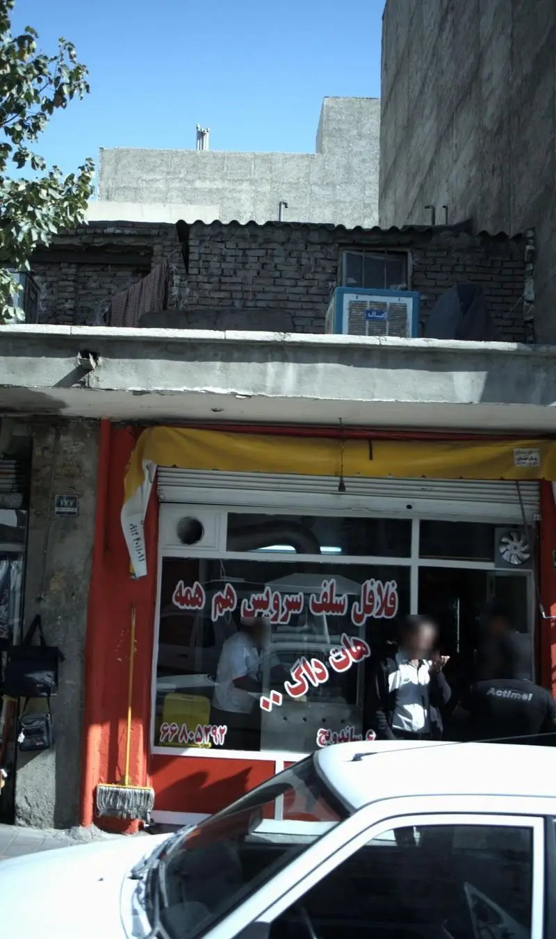 فلافل سلف سرویس هم همه: نمای بیرونی مغازه اغذیه فروشی با مشتریان، ورودی شیشه‌ای، تابلوی زرد و خودروی سفید در خیابان.