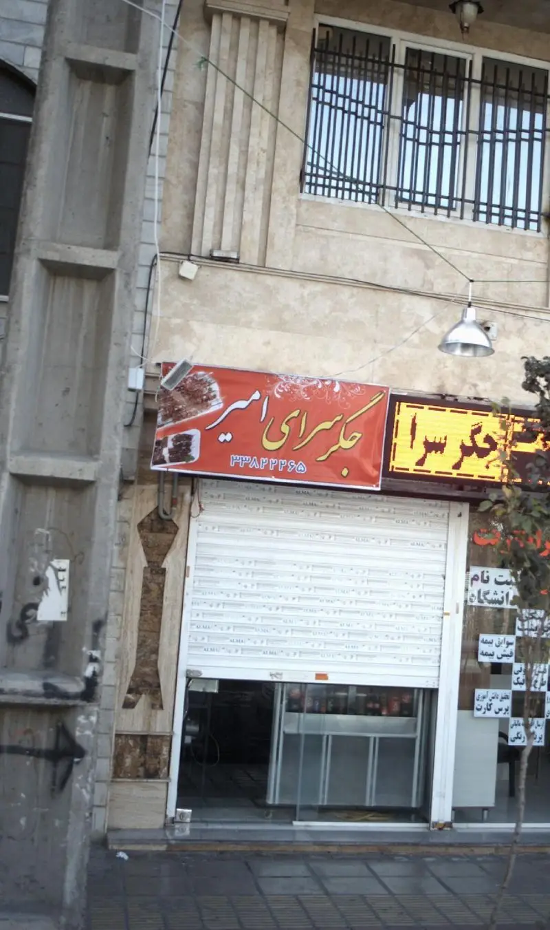 نمای بیرونی جگر سرای امیر با تابلوی قرمز رنگ، کرکره نیمه‌باز و شماره تماس ۳۳۸۲۴۴۶۵ در یک روز آفتابی.