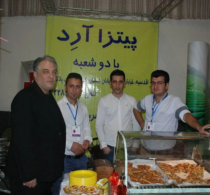 کارکنان پیتزا آرد در یک رویداد معرفی کسب و کار، با تابلوی "پیتزا آرد با دو شعبه" و نمایش انواع پیتزا و غذا.