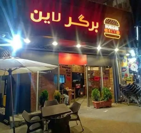 تصویری روشن از نمای بیرونی رستوران برگر لاین در شب با تابلوی نئونی درخشان و لوگوی برگر. این فست فود دارای میز و صندلی در فضای باز است.