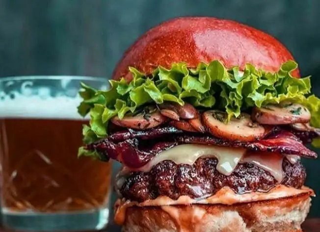 برگر دست‌ساز و لذیذ برگر لاین با نان قرمز، پتی گوشت آبدار، پنیر ذوب‌شده، بیکن ترد، قارچ و کاهو تازه، کنار یک لیوان آبجو خنک.