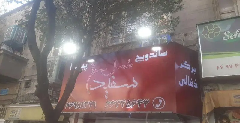 تابلو روشن و دیدنی فست فود سرخ و سفید، ساندویچ و برگر ذغالی با شماره تماس‌های ۹۹۳۳۶۹۶۲۲ و ۹۹۸۱۳۷۷۱ در خیابانی شلوغ.