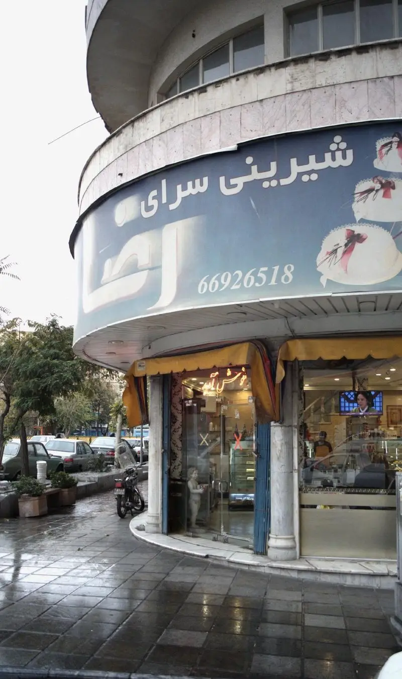 تصویری از نمای بیرونی شیرینی سرای رضا با سردر آبی رنگ، شماره تلفن 66926518 و تصاویر کیک‌ها، پیاده‌رویی خیس و خودروها.
