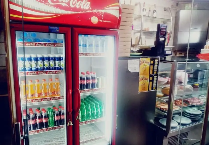 یخچال نوشیدنی بزرگ با برند کوکاکولا در پیتزا گارنی، حاوی انواع نوشابه و آب معدنی. در کنار، ویترین غذاهای آماده و سوسیس.