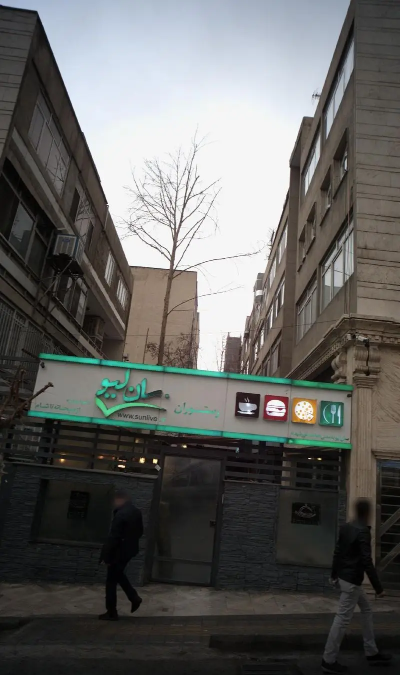نمای بیرونی رستوران سان لایو و طباخی کلپچ خوب در خیابان، با تابلو سبز رنگ و عابران پیاده. ساختمانها و درختان در پسزمینه.