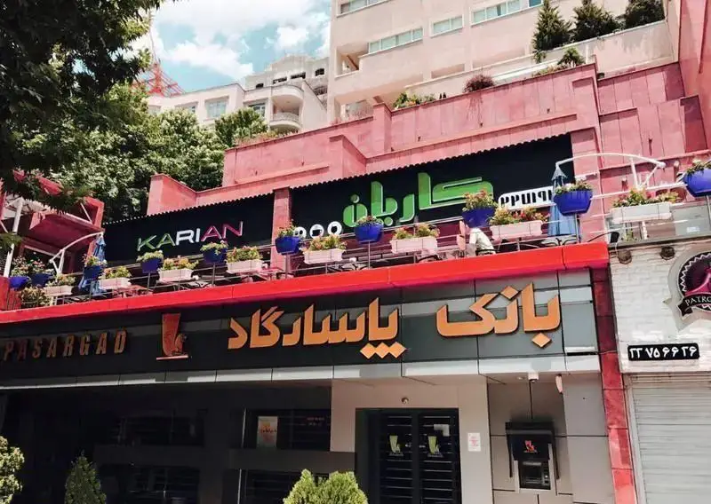 تصویر نمای خارجی رستوران فرنگی کاریان با تابلوی بزرگ و تراس گل‌کاری شده، واقع بر فراز بانک پاسارگاد.