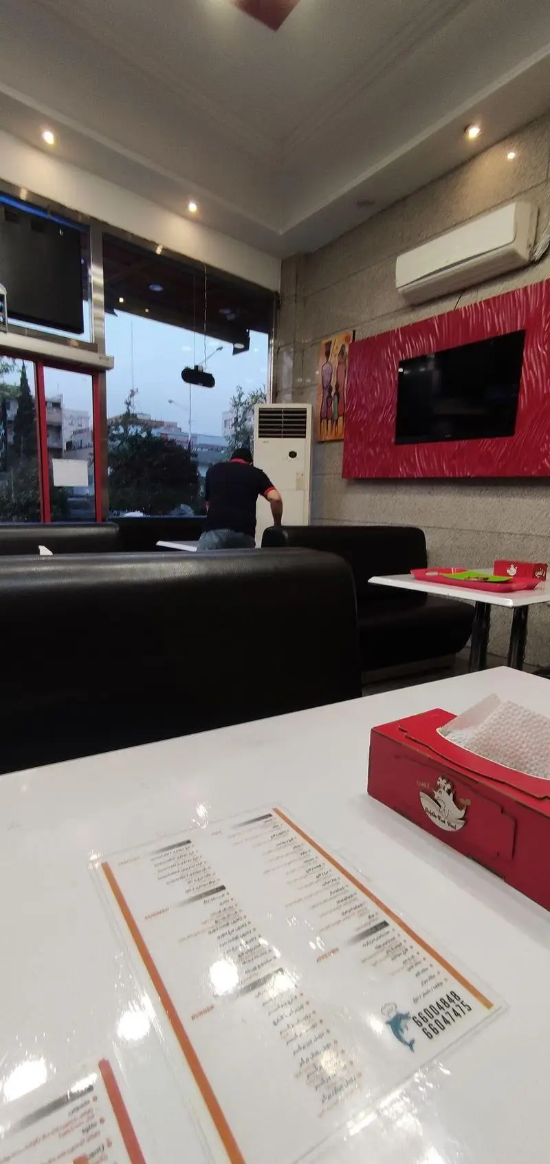 تصویری از داخل فست فود پیتزا دلفین، شامل میز با منو و جعبه دستمال، مبلمان راحت و یک مشتری در محیطی مدرن.