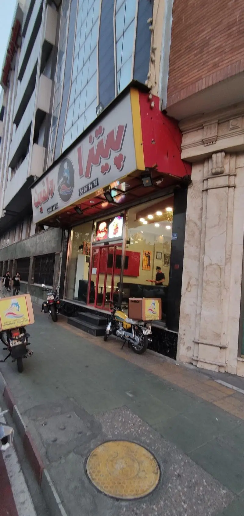 نمای بیرونی فست فود پیتزا دلفین با تابلوی برجسته، دو موتورسیکلت دلیوری و جعبه‌های دلفین در کنار خیابان، آماده پذیرایی از مشتریان.