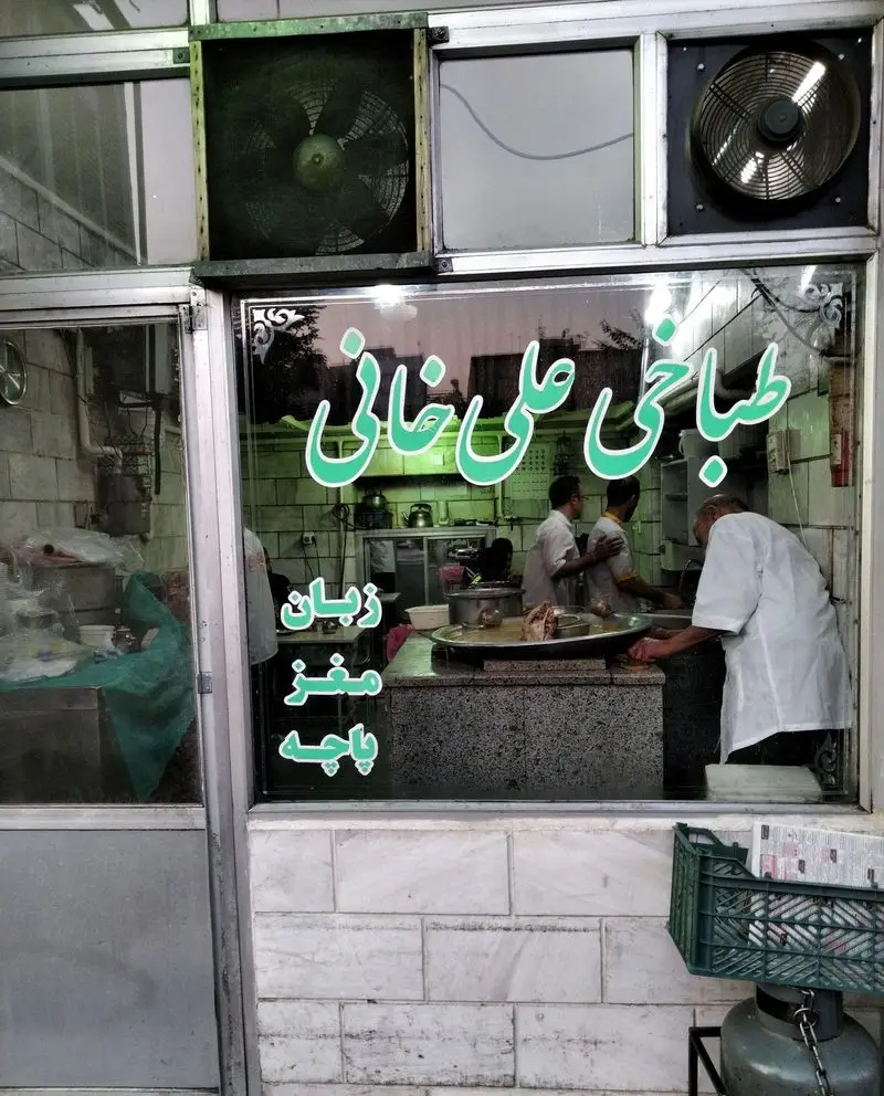 تابلوی طباخی علی خانی با نام و منوی زبان، مغز، پاچه. آشپزها در حال آماده‌سازی کله پاچه سنتی. محیط آشپزخانه و آماده‌سازی غذا دیده می‌شود.