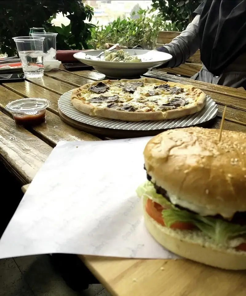 پیتزا گوشت لذیذ و برگر تازه روی میز چوبی رستوران پریماورا، همراه با پاستا و نوشیدنی در فضای باز دلپذیر.