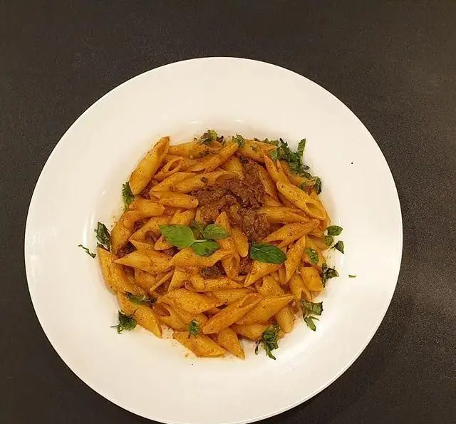 پاستا پنه با سس قرمز و تکه‌های گوشت، تزئین شده با سبزیجات معطر، سرو شده در بشقاب سفید. از منوی خوشمزه پر پل سوخاری.