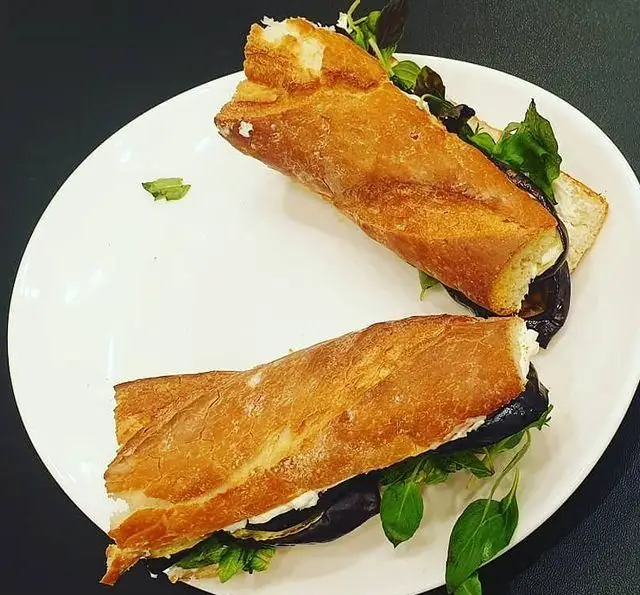 دو نیمه ساندویچ باگت طلایی و لذیذ با مغز بادمجان کبابی، پنیر خامه‌ای و سبزیجات معطر روی بشقاب. از پر پل سوخاری.