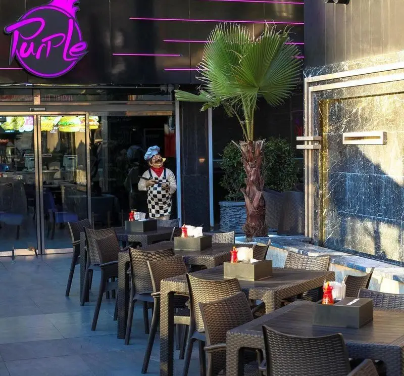 تصویری از نمای بیرونی رستوران فست فود پر پل سوخاری با لوگوی بنفش، فضای نشیمن باز و مجسمه سرآشپز. مکانی ایده‌آل برای صرف غذا.