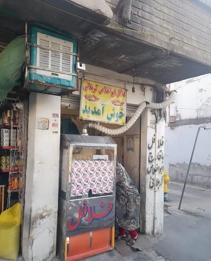 مغازه فلافل ابوالخلاص کربلایی در یک خیابان، با تابلوی "خوش آمدید" و دکه فلافل. کسب و کار محلی کوچک و پر رونق.