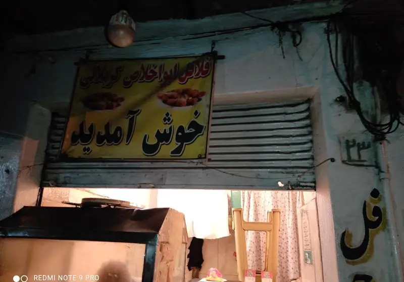 تصویری از ورودی مغازه فلافل‌فروشی «ابو خلاص کربلایی» با تابلوی زرد خوش‌آمدگویی، شامل نام کسب‌وکار و تصاویر فلافل تازه.