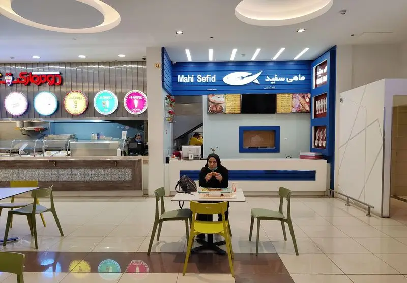 نمای داخلی رستوران ماهی سفید در فودکورت با دکوراسیون آبی و لوگوی ماهی، مشتری نشسته پای میز، منوی غذا و غرفه‌های متنوع دیگر.