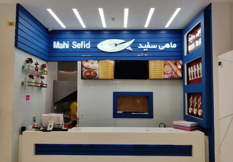 نمای داخلی شیک و آبی-سفید رستوران ماهی سفید، با لوگوی ماهی، میز پذیرش، منوی دیجیتال و بخش آشپزخانه، محیطی دلنشین برای غذای دریایی.