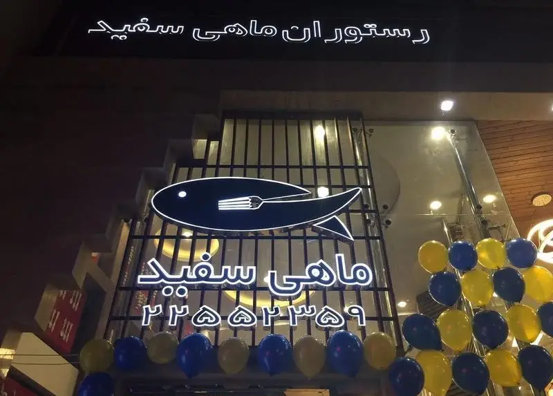 نمای شبانه رستوران ماهی سفید با تابلوی نئونی، لوگوی ماهی و چنگال، شماره تلفن ۲۲۵۵۲۳۵۹ و تزئینات بادکنکی آبی و زرد.