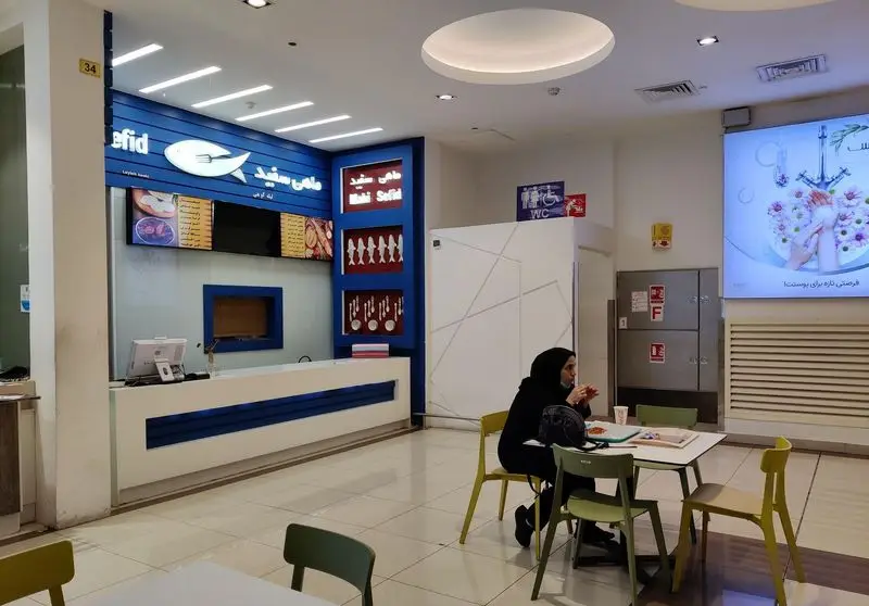 رستوران ماهی سفید: فضای داخلی فودکورت با پیشخوان آبی و سفید، منوی غذاهای دریایی، تزئینات ماهی و مشتریان در حال صرف غذا.