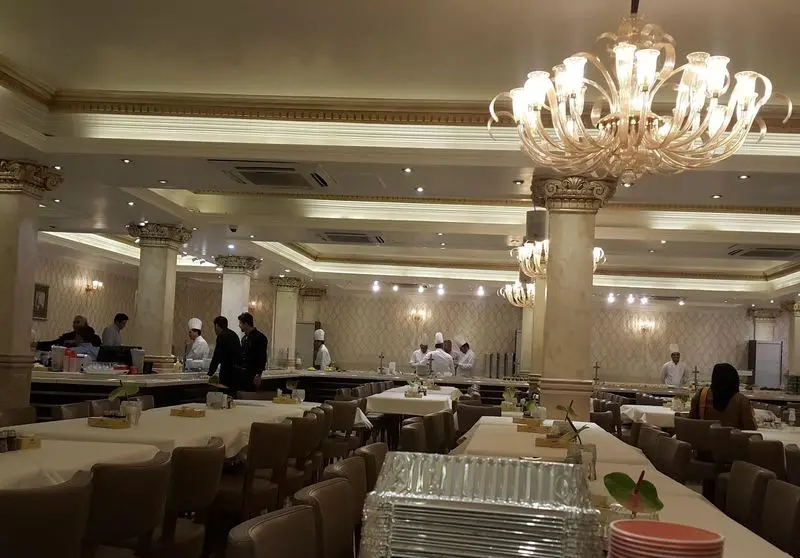 نمای داخلی بزرگ و شیک رستوران هانی مطهری با دکوراسیون لوکس، لوسترهای مجلل، میزهای چیده شده و سرآشپزها در حال کار.