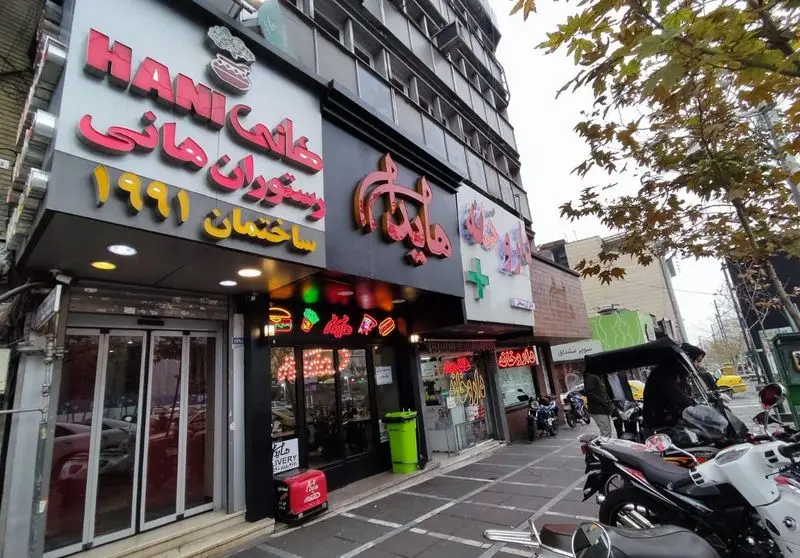 نمای بیرونی رستوران فست فود هانی مطهری با تابلوی "HANI رستوران هانی ۱۹۹۱"، ورودی شیشه‌ای و صندوق دلیوری قرمز رنگ.